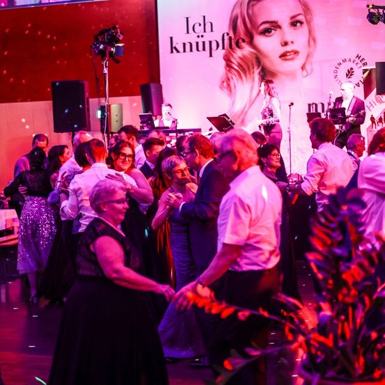 Eine Veranstaltung mit einer großen Bühne, tanzenden Menschen, einer Frau im Rampenlicht und einem rosa Banner im Hintergrund mit der Aufschrift 'Ich knüpfte.'
