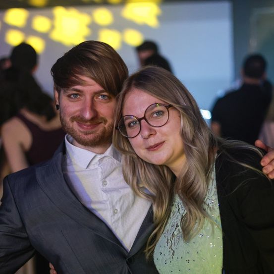 Ein Mann und eine Frau posieren für ein Foto bei einer gesellschaftlichen Veranstaltung, lächeln warm und haben andere im Hintergrund.