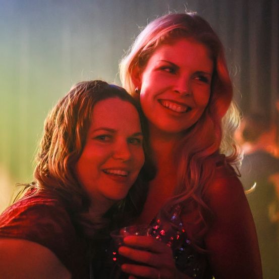 Zwei Frauen lächeln und posieren für ein Foto auf einer Party. Sie halten Getränke in den Händen.