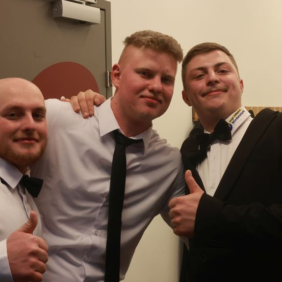 Drei Männer posieren für ein Foto. Der mittlere Mann trägt ein weißes Hemd und eine Krawatte, während die anderen schwarze Anzüge und Krawatten tragen. Sie lächeln und zeigen Daumen hoch.