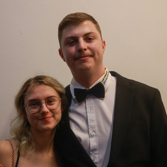 Ein Mann und eine Frau posieren für ein Foto. Der Mann trägt einen Smoking mit Fliege und eine Namensplakette mit der Aufschrift 'Blindermann'. Die Frau trägt eine Brille und ein trägerloses Kleid. Sie stehen eng beieinander und scheinen zu lächeln.
