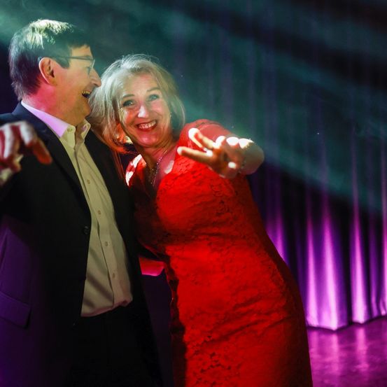 Ein Mann und eine Frau tanzen auf einer Bühne mit lila Lichtern. Die Frau trägt ein rotes Kleid und lächelt. Der Mann trägt einen Anzug und zeigt auf etwas.