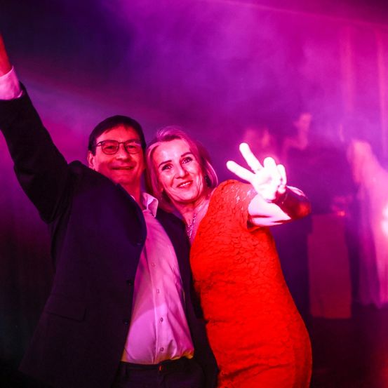 Ein Mann und eine Frau tanzen bei einer Party mit lila Lichtern. Der Mann hebt seine linke Hand, während die Frau ein Friedenszeichen macht.