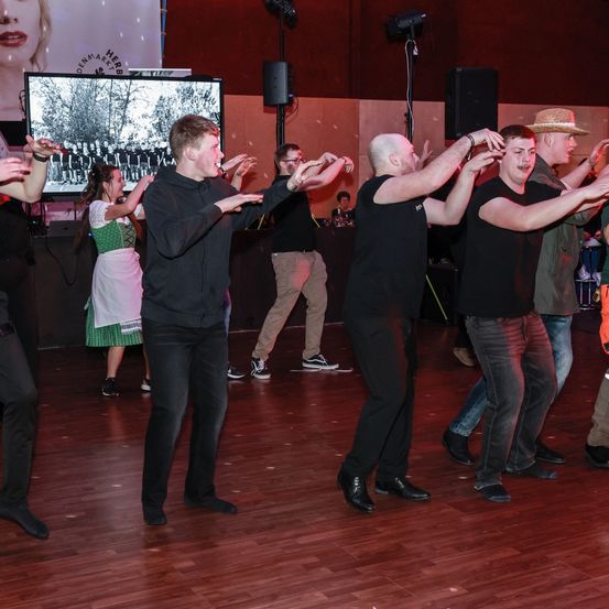 Men tanzen auf einem Holzboden, einer trägt einen Hut. Eine Frau in einem grünen Kleid ist in der Nähe. Lautsprecher und ein großer Bildschirm, der ein Gruppenfoto zeigt, sind im Hintergrund.