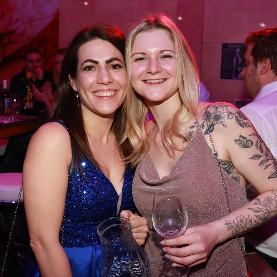 Zwei lächelnde Frauen bei einer Veranstaltung, eine in Blau und eine in Pink, halten Getränke. Sie sind von verschwommenen Menschen umgeben.
