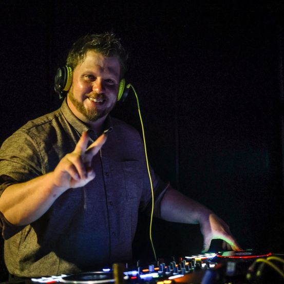Ein lächelnder Mann mit Kopfhörern steht in einem schwach beleuchteten Raum und zeigt mit einer Hand. Vor ihm steht ein DJ-Mixer mit bunten Lichtern.