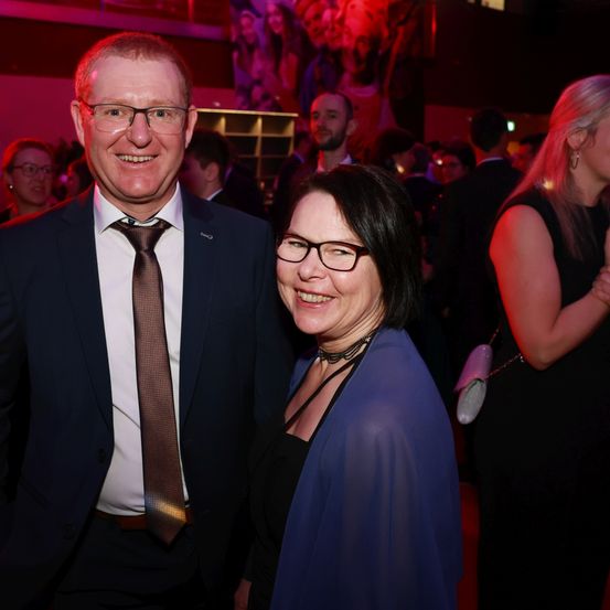 Ein Mann und eine Frau lächeln und stehen bei einer formellen Veranstaltung eng beieinander. Sie tragen beide eine Brille und sind formell gekleidet. Hinter ihnen stehen ein paar Leute und scheinen zu sprechen. Der Hintergrund ist unscharf, mit einem roten Licht, das den Bereich beleuchtet.