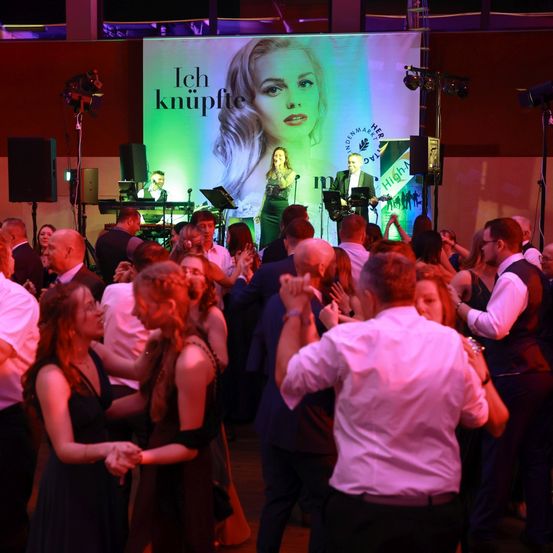 Eine Menschenmenge tanzt auf einer Bühne, vor der ein großer Bildschirm im Hintergrund hängt. Der Bildschirm zeigt das Gesicht einer Frau mit den Worten 'Ich knüpfie'. Vor dem Bildschirm spielt eine Band.