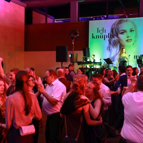 Eine Veranstaltung mit Bühne, einer großen Projektion einer Frau und einer Live-Band. Das Publikum tanzt und genießt die Musik.