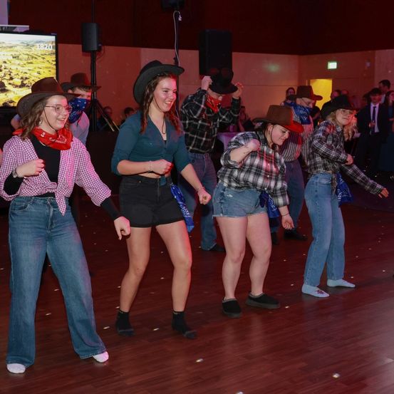 Eine Gruppe von Frauen in Western-Outfits tanzt auf einer Bühne. Sie tragen Cowboy-Hüte, karierte Hemden und Jeansshorts. Eine Frau trägt eine Brille und ein rotes Halstuch. Die Bühne hat einen Holzboden und ist schwach beleuchtet.