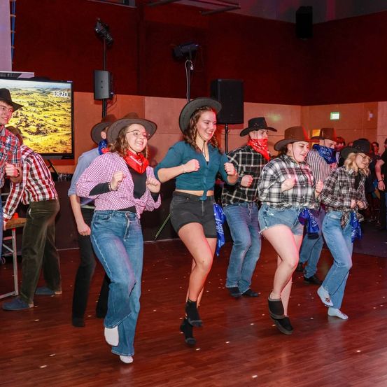 Eine Gruppe von Menschen in Cowboyhüten und Stiefeln tanzt auf einer hölzernen Tanzfläche. Sie befinden sich in einem Raum mit einer großen Leinwand im Hintergrund.