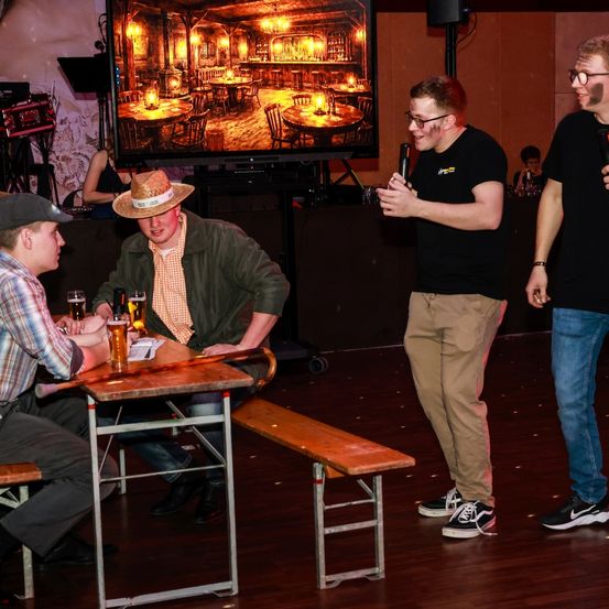 Eine Gruppe junger Männer sitzt an einem Tisch in einem schwach beleuchteten Raum, mit einem großen Bildschirm, der eine Barszene hinter ihnen zeigt. Ein Mann hält ein Mikrofon, während ein anderer ein Bierglas hält. Eine Bank steht in der Nähe.