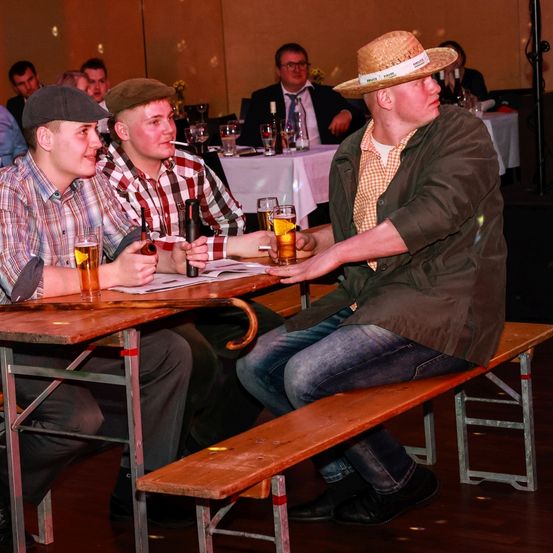 Mehrere junge Männer sitzen in einer Bar an einem Tisch, einer von ihnen raucht. Sie halten Gläser Bier in den Händen und einer von ihnen gibt eine High Five. Hinter ihnen steht ein Mann im Anzug.