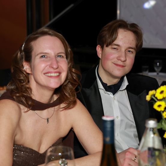 Eine Frau mit lockigem Haar und einer Halskette sitzt neben einem Mann mit schwarzer Fliege, beide lächeln in die Kamera. Sie sitzen an einem Tisch mit Weingläsern und einer Sonnenblumen-Arrangement.