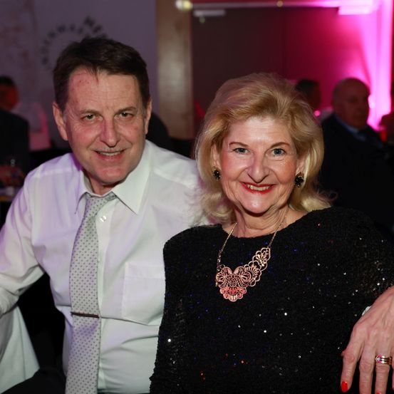Ein Mann und eine Frau lächeln für ein Foto bei einer formellen Veranstaltung, mit rosa Beleuchtung im Hintergrund.