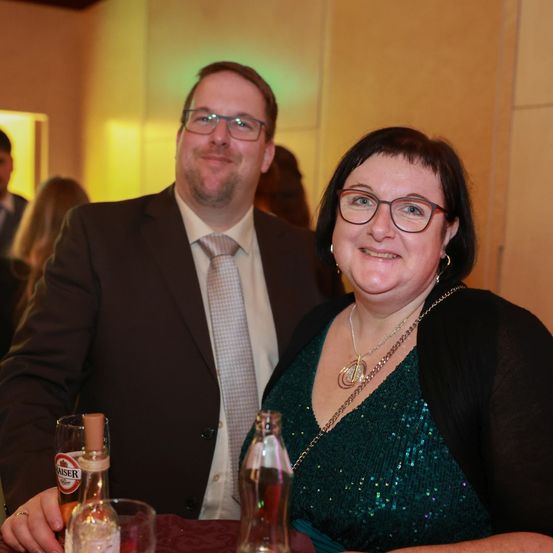 Ein Mann und eine Frau lächeln und posieren für ein Foto. Sie sind bei einer formellen Veranstaltung und sitzen an einem Tisch mit Getränken.