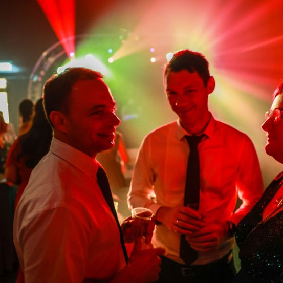 Drei Personen befinden sich in einem Partyraum. Zwei Männer und eine Frau lächeln und halten Getränke in den Händen. Der Raum ist von roten und grünen Lichtern beleuchtet.