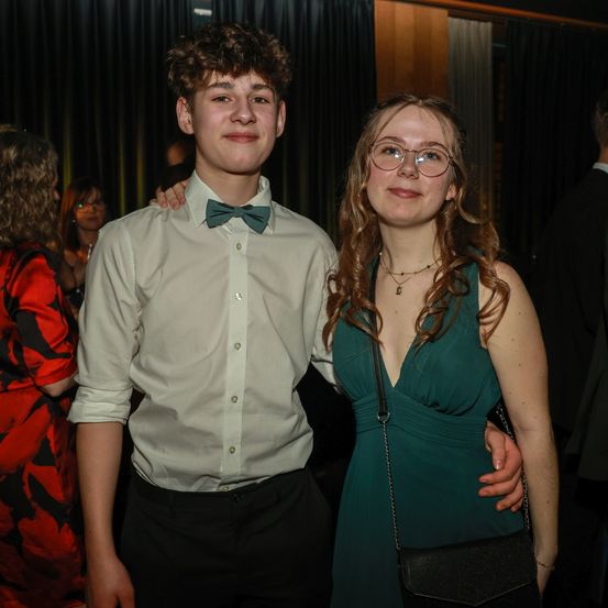 Ein junges Paar steht eng beieinander bei einer formellen Veranstaltung. Der Mann trägt ein helles Hemd und eine Fliege, während die Frau lange lockige Haare, eine Brille und ein grünes Kleid hat. Sie lächeln und scheinen für ein Foto zu posieren.