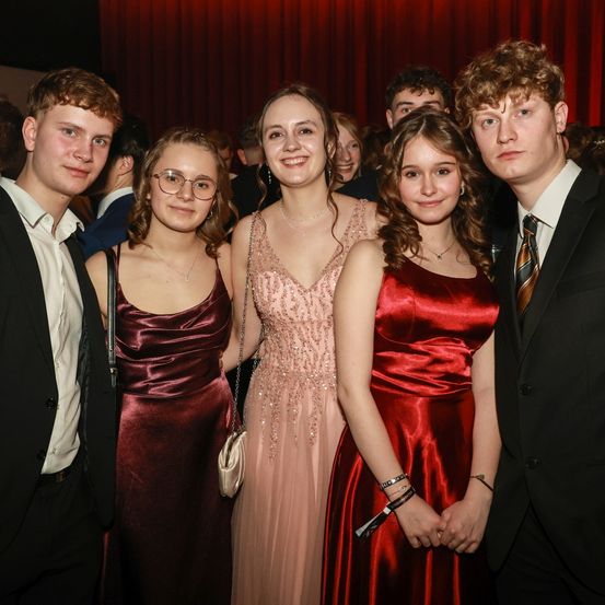 Eine Gruppe junger Leute in formeller Kleidung posiert für ein Foto bei einer Veranstaltung. Drei Frauen und zwei Männer lächeln und stehen eng beieinander. Ein roter Vorhang ist hinter ihnen.