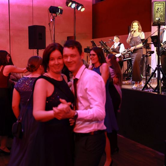Eine Gruppe von Menschen tanzt auf einer Bühne, die von rosa Lichtern beleuchtet wird, während eine Frau singt und eine Band im Hintergrund spielt.
