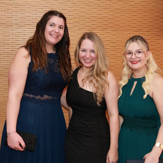 Drei Frauen in Abendkleidern stehen eng beieinander und lächeln für ein Foto vor einer braunen Wand. Die linke Frau hält eine schwarze Handtasche, während die mittlere und die rechte eine Brille tragen.