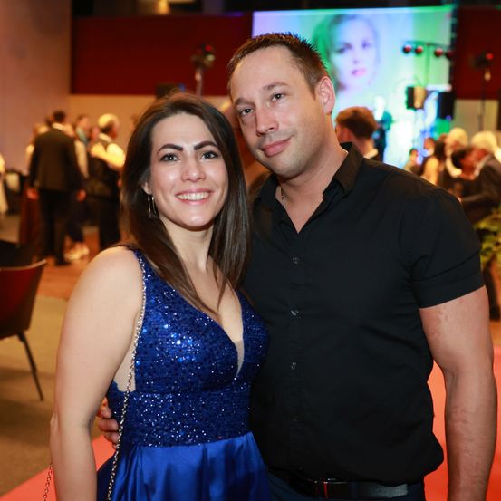 Ein lächelndes Paar bei einer Veranstaltung, die Frau in einem blauen Kleid und der Mann in einem schwarzen Hemd, stehen auf einem roten Teppich mit Menschen im Hintergrund.