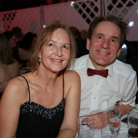 Ein Paar, das für ein Foto bei einer Veranstaltung lächelt. Die Frau trägt ein schwarzes Kleid und der Mann ein weißes Hemd mit roter Schleife. Hinter ihnen sind verschwommene Figuren von sitzenden und stehenden Personen zu sehen, mit einem rosa Zaun im Hintergrund.