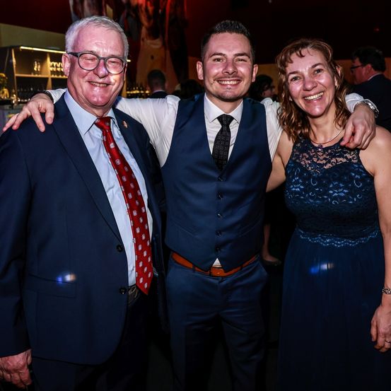 Drei Personen lächeln für ein Foto bei einer Veranstaltung; ein Mann mit Brille und roter Krawatte, ein Mann in einem blauen Vest und eine Frau in einem blauen Kleid.