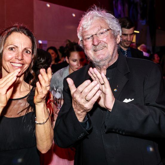 Ein Mann und eine Frau lächeln und klatschen bei einer Veranstaltung. Der Mann trägt eine Brille und einen schwarzen Anzug. Die Frau trägt ein schwarzes Kleid und eine Armband.