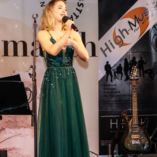 Eine Frau in einem grünen Kleid steht auf der Bühne und hält ein Mikrofon. Hinter ihr ist eine Gitarre prominent zu sehen. Ein Banner mit der Aufschrift 'High-Music' ist sichtbar.
