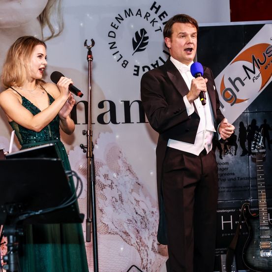 Ein Mann und eine Frau auf der Bühne, die Frau hält ein Mikrofon, der Mann singt in sein. Ein Musikbanner und eine Gitarre im Hintergrund.