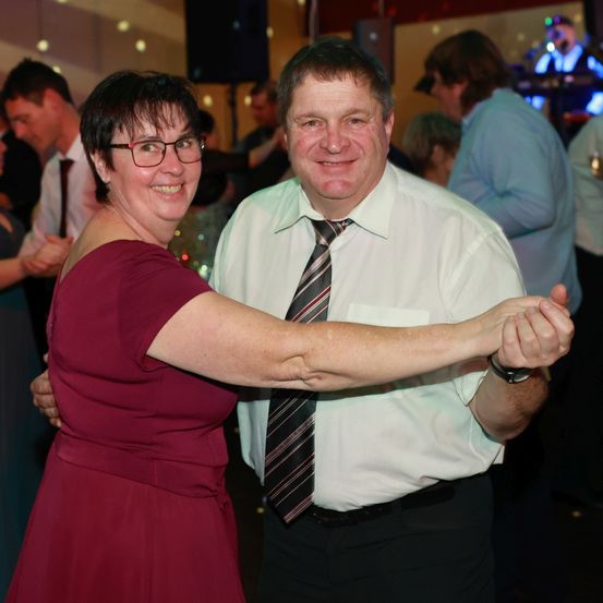 Ein Mann und eine Frau tanzen in einem überfüllten Raum. Der Mann trägt ein weißes Hemd und eine Krawatte. Die Frau trägt ein rotes Kleid und eine Brille. Andere Leute tanzen und stehen hinter ihnen.