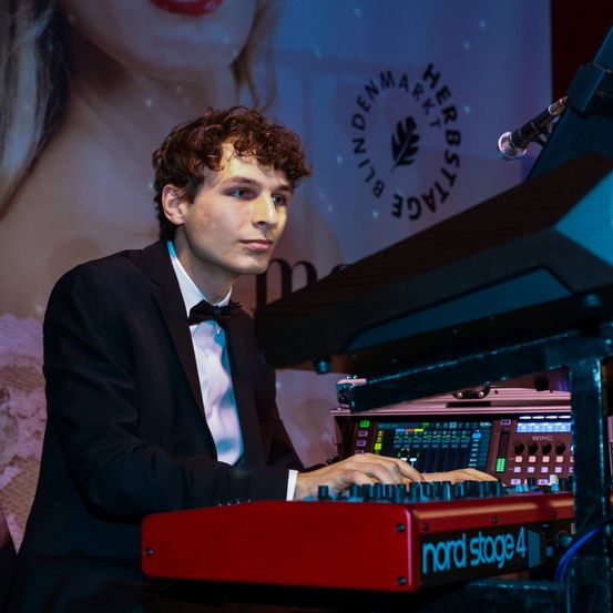 Ein Mann in Anzug und Fliege spielt an einem roten Keyboard, während ein Mikrofon über ihm positioniert ist. Im Hintergrund ist eine Frau in einem rosa Kleid zu sehen.