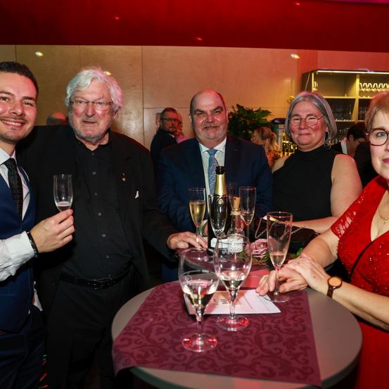 Eine Gruppe von Menschen in formeller Kleidung posiert für ein Foto um einen Tisch mit Weingläsern und einer Flasche Champagner.