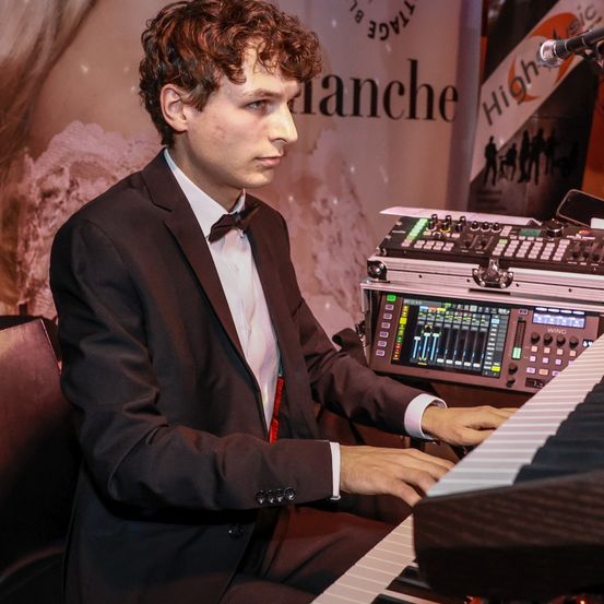 Ein Mann in Anzug und Fliege sitzt an einem Klavier und spielt konzentriert. Hinter ihm steht ein großes Schild mit der Aufschrift 'Blanche' und ein Musiksteuerpult befindet sich auf dem Tisch.