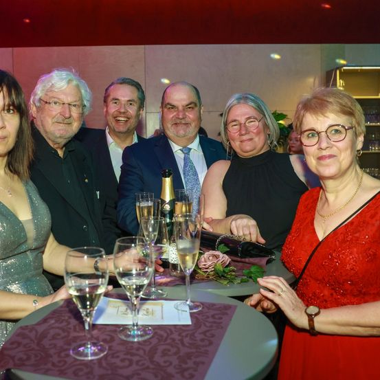 Sechs Personen, vier Männer und zwei Frauen, posieren für ein Foto bei einer Veranstaltung. Sie lächeln und einige halten Weingläser. Sie stehen vor einem Tisch mit einer Champagnerflasche.