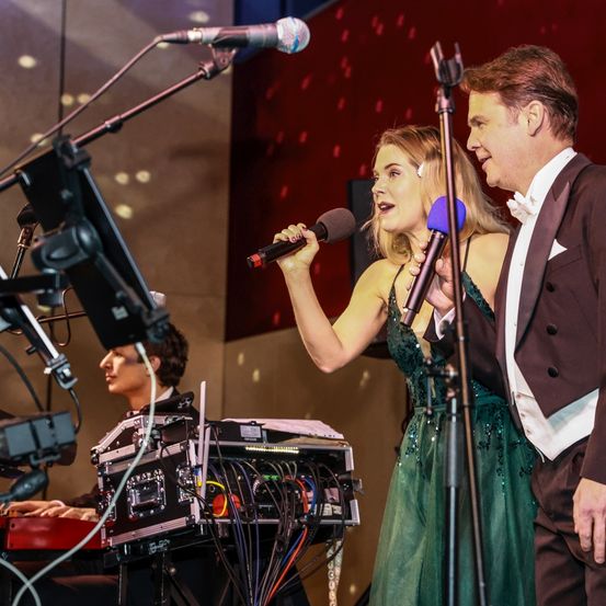 Eine Frau in einem grünen Kleid singt in ein Mikrofon, begleitet von einem Mann im Frack. Hinter ihnen spielt ein Pianist auf einem Keyboard. Ausrüstung wie Mikrofone, Lautsprecher und Kameras sind auf der Bühne aufgebaut.