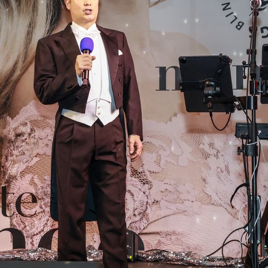 Ein Mann in einem dunklen Anzug hält ein violettes Mikrofon und steht auf der Bühne mit einem Notenständer und Kabeln dahinter. Der Hintergrund zeigt ein rosa Spitzendesign.