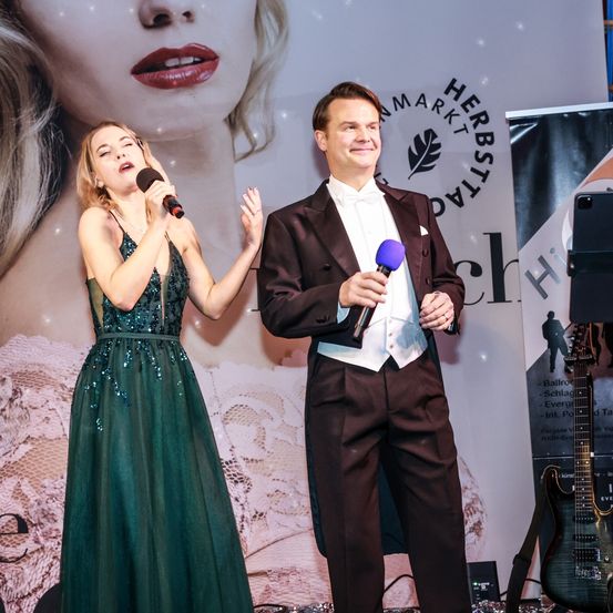 Eine Frau in einem grünen Kleid und ein Mann in einem schwarzen Anzug stehen auf der Bühne mit Mikrofonen und singen. Dahinter ist ein großes Bild des Gesichts einer Frau und ein Banner mit Text.
