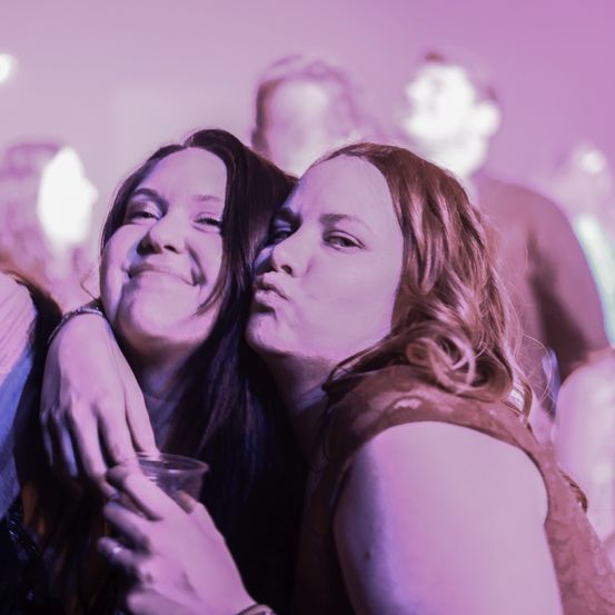 Zwei Frauen stehen eng beieinander, lächeln und halten Händchen. Sie sind wahrscheinlich bei einem Konzert, und die Bühne ist mit einem lila Licht beleuchtet.