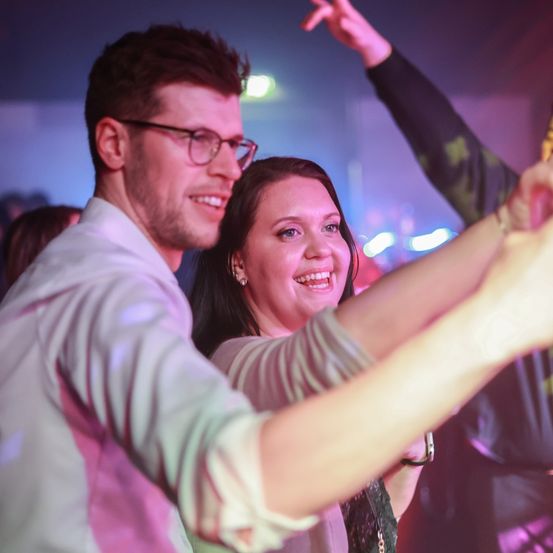 Ein lächelnder Mann und eine Frau tanzen in einem Nachtclub, beleuchtet von lila Lichtern.