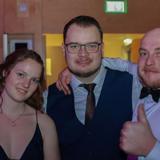 Drei Menschen posieren für ein Foto; eine Frau in einem schwarzen Kleid, ein Mann in einem blauen Weste und Krawatte und ein weiterer Mann mit Fliege, der einen Daumen hochhält.