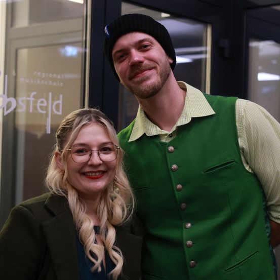 Ein Mann und eine Frau stehen lächelnd zusammen draußen. Der Mann trägt eine grüne Weste und eine schwarze Mütze. Die Frau trägt eine Brille und eine grüne Jacke. Sie stehen vor einer Glastür mit weißer Schrift.
