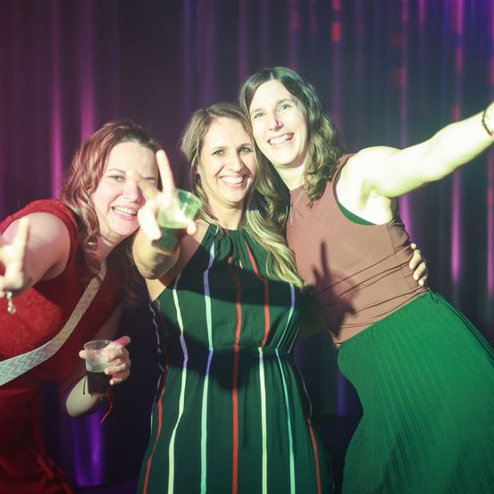 Drei Frauen lächeln und posieren für ein Foto auf einer Party. Die Frau links macht ein Friedenszeichen und hält ein Getränk. Die mittlere Frau hält ein Glas. Die Frau rechts hat ihren Arm um die mittlere Frau gelegt.