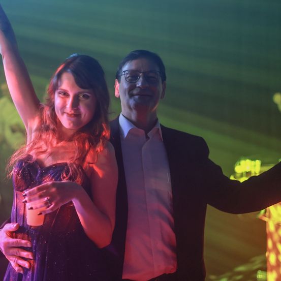 Eine Frau und ein Mann stehen zusammen mit umschlungenen Armen auf einer Party. Die Frau lächelt und der Mann trägt eine Brille. Sie halten Getränke in den Händen. Hinter ihnen ist ein grüner Hintergrund.