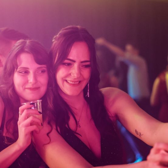 Zwei Frauen lächeln und posieren für ein Foto in einem schwach beleuchteten Raum, eine hält ein Getränk, während andere im Hintergrund tanzen.