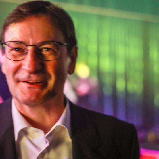 Ein lächelnder Mann mit Brille und Anzug steht vor einem verschwommenen Hintergrund mit grünen und lila Lichtern.