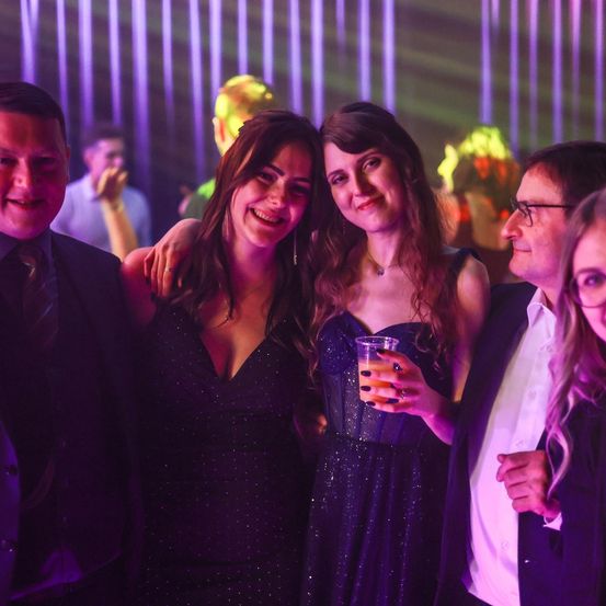 Zwei Frauen und zwei Männer stehen in einem Club und lächeln. Die Frauen tragen Abendkleider. Der Mann rechts hält ein Getränk. Der Hintergrund ist lila und grün.