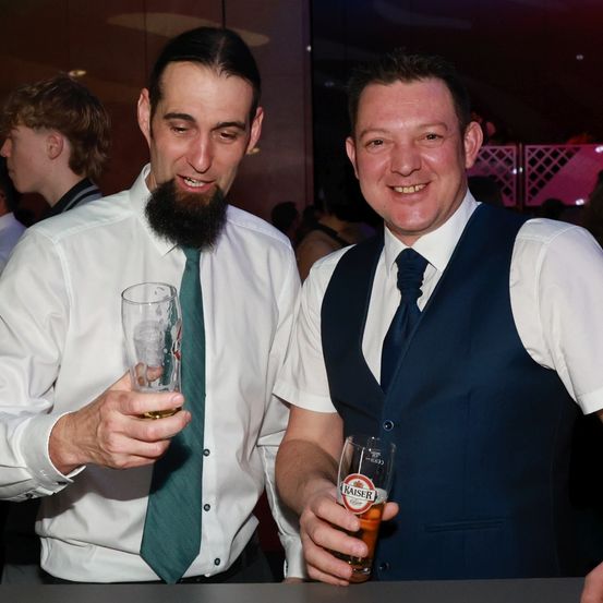 Zwei Männer auf einer Party, einer mit Bart und grüner Krawatte, hält ein Glas, der andere in einem Weste, hält ein Glas Kaiser Bier. Sie lächeln, mit Leuten im Hintergrund.