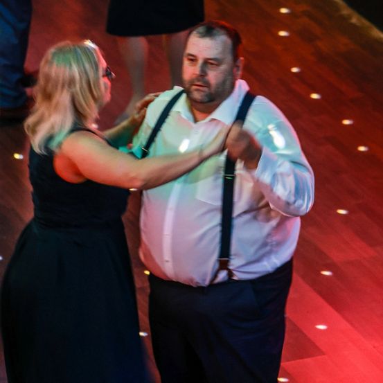 Ein Mann und eine Frau tanzen auf einer roten Tanzfläche, wobei der Mann eine Hosenträger trägt und die Frau ein schwarzes Kleid.